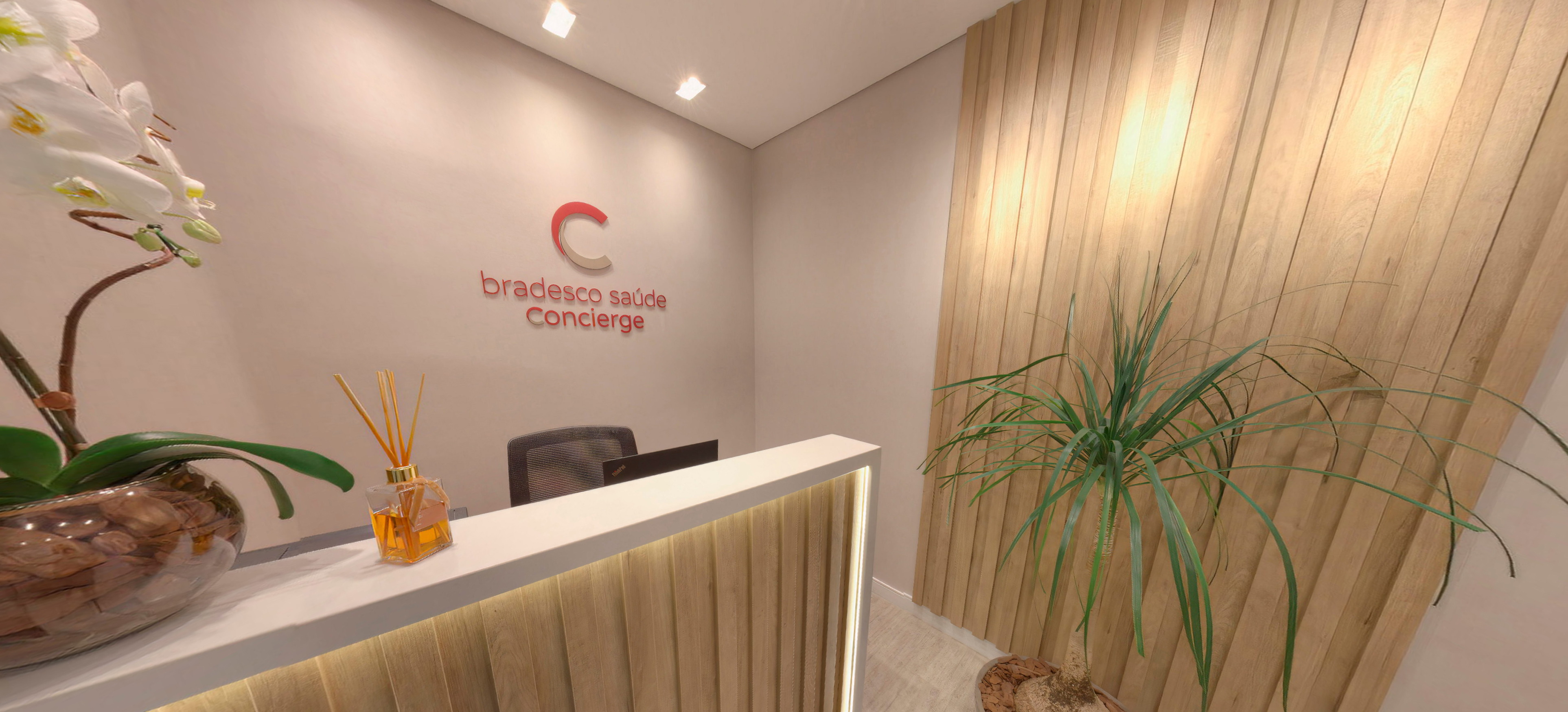 Sala de atendimento, com mesa de vidro, computador e cadeira de escritório. Ao lado da mesa, há um vaso decorativo de orquídeas.