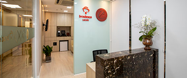 Sala de recepção com balcão em granito, com vaso decorativo e cadeira de escritório atrás do balcão. Ao fundo, há a sala de atendimento com poltrona de couro, na cor bege, televisão, frigobar e cafeteira expressa.